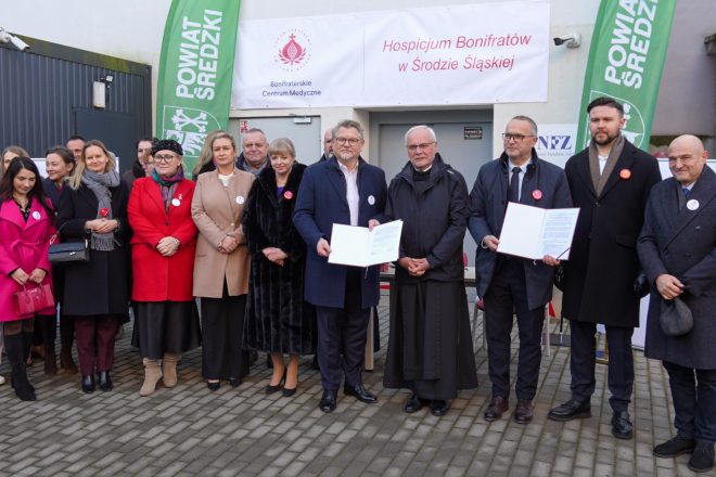 Duża grupa uczestników uroczystości stoi przed wejściem do nowo otwartego hospicjum Bonifratrów w Środzie Śląskiej. Na środku, w pierwszym rzędzie, czterej mężczyźni prezentują otwarte teczki z podpisanymi dokumentami. Po obu stronach stoją przedstawiciele lokalnych instytucji, personel oraz osoby duchowne. W tle widoczne są banery Bonifraterskiego Centrum Medycznego i Powiatu Średzkiego