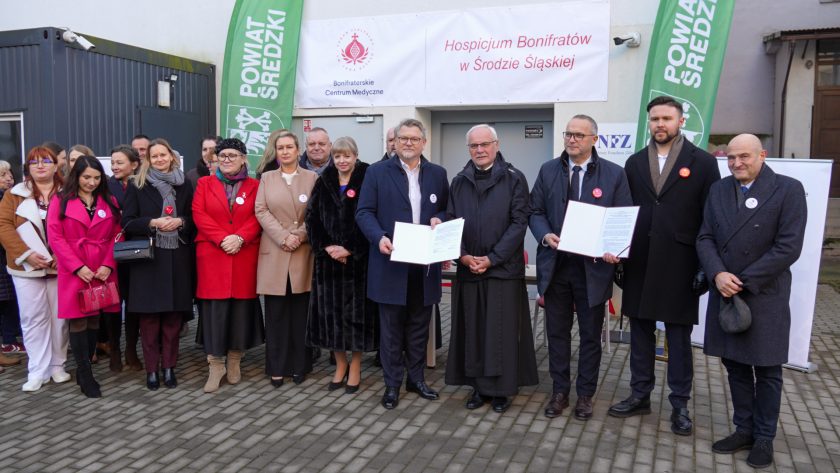 Duża grupa uczestników uroczystości stoi przed wejściem do nowo otwartego hospicjum Bonifratrów w Środzie Śląskiej. Na środku, w pierwszym rzędzie, czterej mężczyźni prezentują otwarte teczki z podpisanymi dokumentami. Po obu stronach stoją przedstawiciele lokalnych instytucji, personel oraz osoby duchowne. W tle widoczne są banery Bonifraterskiego Centrum Medycznego i Powiatu Średzkiego