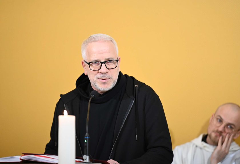 Zakonnik o siwych włosach i w okularach, ubrany w czarny habit i bluzę z kapturem, stoi przy ambonie przed mikrofonem i czyta z księgi liturgicznej. Obok widoczna jest zapalona świeca. W tle siedzi inny zakonnik w białym habicie