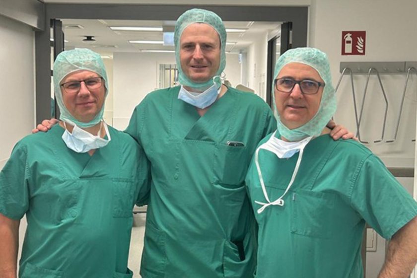 Trzech lekarzy w zielonych strojach i czepkach ochronnych pozuje razem na szpitalnym korytarzu, stojąc ramię w ramię i uśmiechając się do zdjęcia