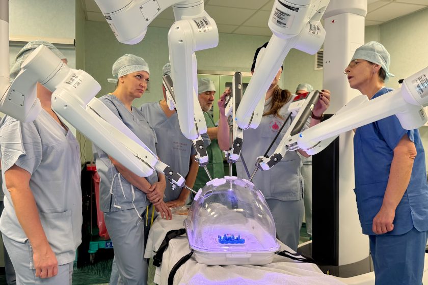 Grupa pielęgniarek i personelu bloku operacyjnego w strojach chirurgicznych obserwuje pracę ramion robota da Vinci podczas szkolenia. Uczestnicy stoją wokół trenażera umieszczonego pod przezroczystą kopułą, analizując ruchy robota