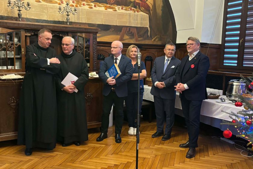 Uczestnicy spotkania opłatkowego stojący w półokręgu w zabytkowej sali, w centrum mikrofon, w tle świąteczna dekoracja