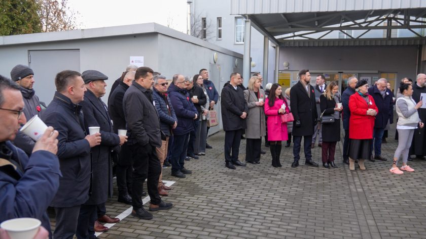 Szerokie ujęcie dużej grupy uczestników zgromadzonych na placu przed budynkiem. Wszyscy stoją w półkolu, patrząc w kierunku przemawiających. Wiele osób trzyma papierowe kubki z gorącym napojem. W tle widoczne są drzwi i ściany budynku oraz zadaszenie nad wejściem