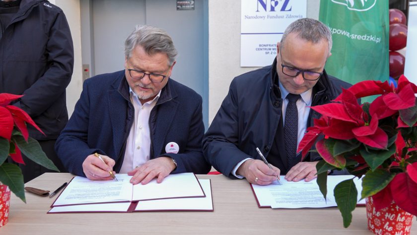 Dwaj elegancko ubrani mężczyźni siedzą przy stole na zewnątrz budynku i podpisują oficjalne dokumenty umieszczone w bordowych teczkach. Obaj koncentrują się na podpisywaniu, trzymając pióra w dłoniach. Na pierwszym planie po obu stronach stołu stoją czerwone poinsecje. W tle widoczne są tablice informacyjne Narodowego Funduszu Zdrowia oraz roll-up Powiatu Średzkiego