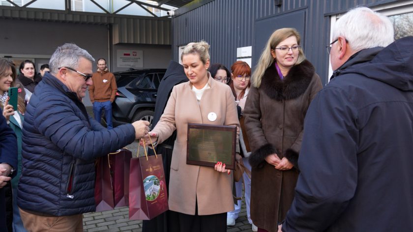 Przed budynkiem hospicjum mężczyzna w granatowej kurtce wręcza prezenty w bordowych torbach. Kobieta w beżowym płaszczu przyjmuje upominek, trzymając jednocześnie dużą, złotą tablicę – certyfikat akredytacyjny. Obok niej stoi kobieta w brązowym płaszczu z futrzanym kołnierzem. W tle zgromadzeni uczestnicy obserwują moment wręczenia