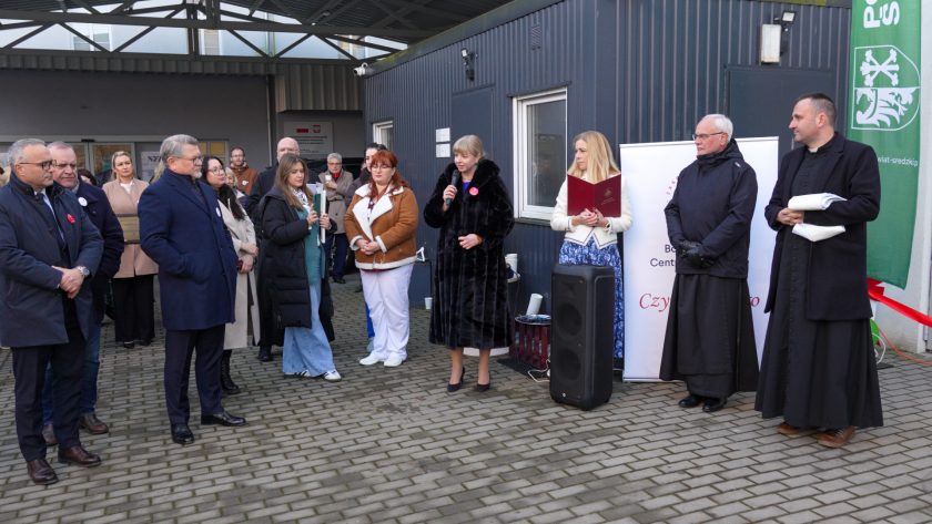 Grupa osób zgromadzona na dziedzińcu słucha przemówienia kobiety w czarnym futrzanym płaszczu, mówiącej do mikrofonu. Obok niej stoi kobieta z bordową teczką, a za nimi widoczni są zakonnicy oraz roll-up Bonifraterskiego Centrum Medycznego. Zebrani goście ustawieni są półkolem, część trzyma kubki z gorącym napojem