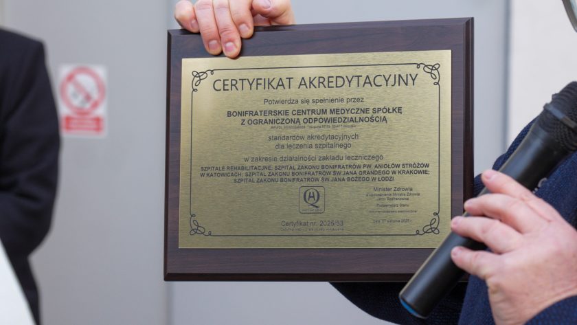 Ujęcie przedstawia dłonie osoby trzymającej elegancką, złotą tablicę z napisem „Certyfikat Akredytacyjny” przyznany Bonifraterskiemu Centrum Medycznemu sp. z o.o. Tablica znajduje się w drewnianej ramie, a obok widoczny jest fragment mikrofonu