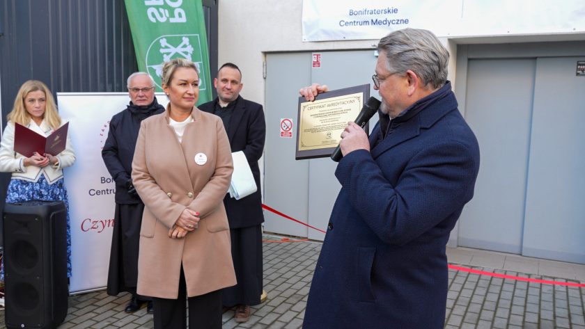 Mężczyzna w granatowym płaszczu trzyma certyfikat akredytacyjny i prezentuje go publiczności, mówiąc do mikrofonu. Przed nim stoi kobieta w beżowym płaszczu, a za nią dwaj zakonnicy i prowadząca z teczką. W tle widoczny jest baner hospicjum i zielony roll-up Powiatu Średzkiego