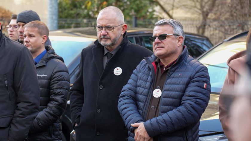 Dwóch mężczyzn stoi obok siebie, słuchając przemówienia. Jeden w czarnym eleganckim płaszczu z przypiętą odznaką, drugi w granatowej pikowanej kurtce i okularach przeciwsłonecznych. W tle widoczni są inni uczestnicy oraz zaparkowane samochody
