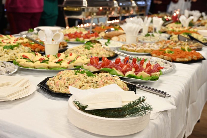 Zbliżenie stołu z uroczystym poczęstunkiem – półmiski z sałatkami i przekąskami, kanapki z dekoracją z pomidorków, jednorazowe sztućce i talerzyki; na pierwszym planie stos białych talerzy ozdobiony gałązką świerku