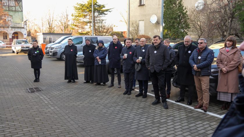 Szersze ujęcie grupy ludzi stojących na zewnątrz podczas ceremonii. Wśród nich znajdują się zakonnicy, przedstawiciele instytucji oraz goście, część trzyma papierowe kubki. W tle widoczne są budynki, samochody oraz zimowa roślinność