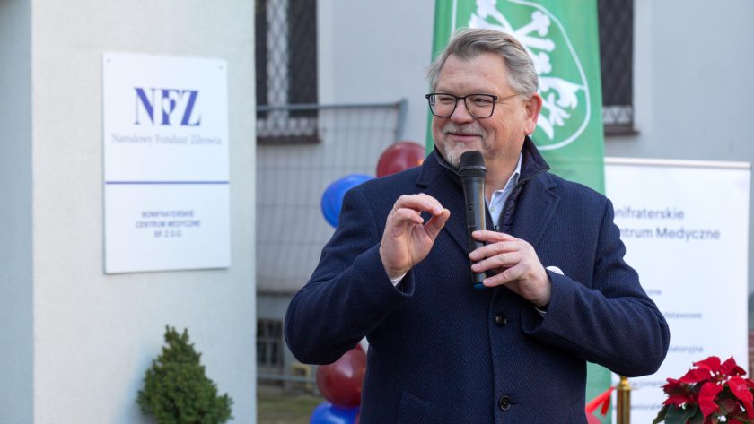 Mężczyzna w granatowym płaszczu przemawia do mikrofonu, gestykulując ręką. W tle widać tablicę Narodowego Funduszu Zdrowia, zieloną flagę Powiatu Średzkiego oraz balonowe dekoracje w barwach czerwieni i błękitu