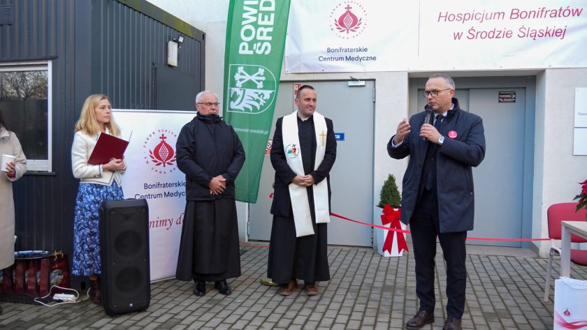 Mężczyzna w eleganckim płaszczu i okrągłym czerwonym znaczku mówi do mikrofonu na tle wejścia do hospicjum. Po jego lewej stronie stoją kobieta z teczką, zakonnik oraz kapłan w stule. Za nimi widoczny jest roll-up Bonifraterskiego Centrum Medycznego
