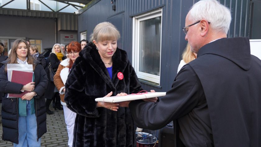 Kobieta w czarnym futrzanym płaszczu odbiera dokument od zakonnika w czarnym habicie. W tle widać pozostałych uczestników uroczystości, w tym kobietę trzymającą teczkę i kilka osób z uśmiechem obserwujących sytuację