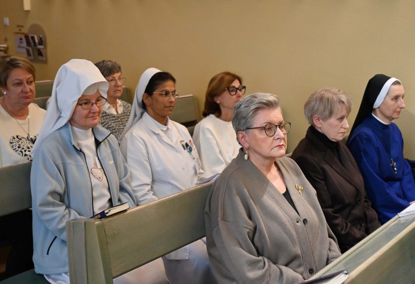 Kilka kobiet siedzi w ławce kaplicy, w tym siostry zakonne w białych i granatowych habitach oraz kobiety świeckie. Wszystkie są skupione na modlitwie lub słuchaniu. Przed jedną z sióstr leży książeczka do nabożeństwa