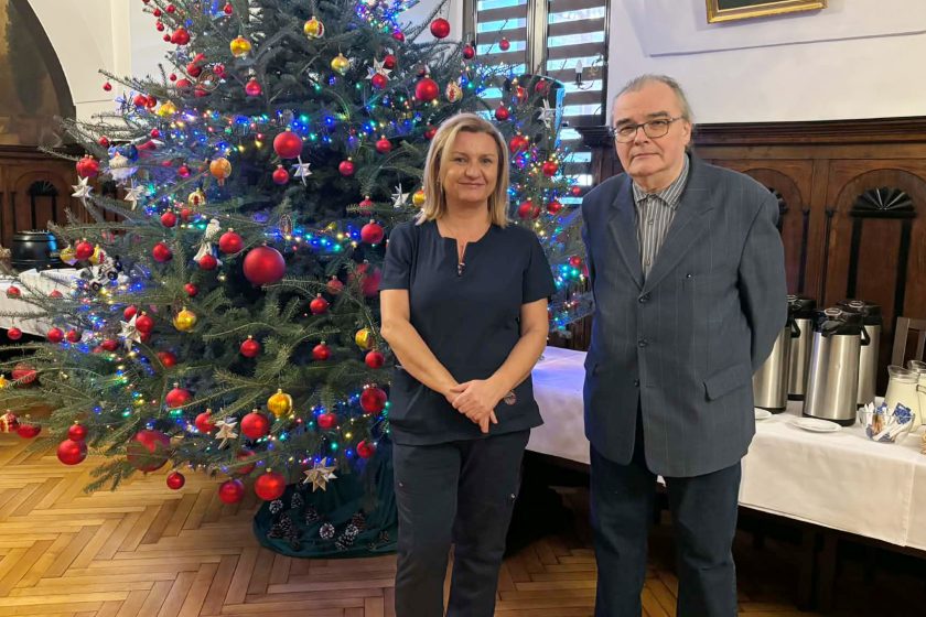 Dwóch uczestników spotkania opłatkowego pozujących do zdjęcia na tle udekorowanej choinki bożonarodzeniowej