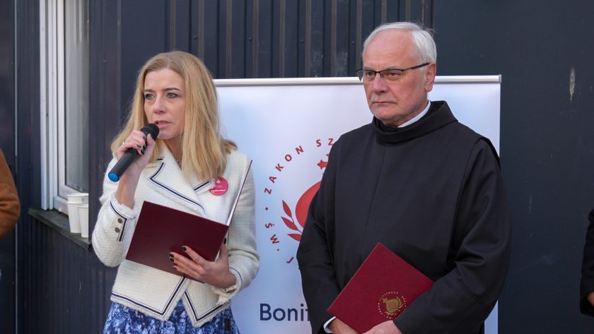 Kobieta w białym żakiecie i niebieskiej spódnicy mówi do mikrofonu, trzymając bordową teczkę. Obok niej stoi brat zakonny w czarnym habicie, trzymający podobną teczkę i patrzący prosto przed siebie. Za nimi znajduje się roll-up z logo Zakonu Bonifratrów