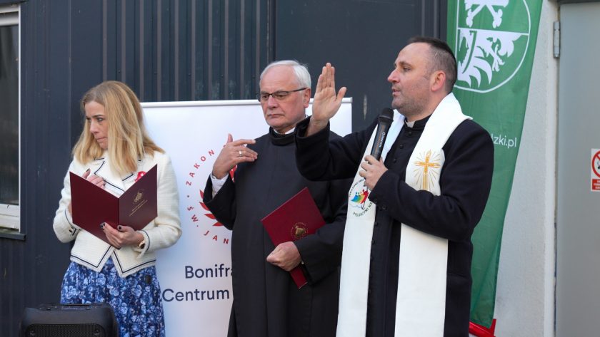 Trzy osoby stoją obok siebie podczas uroczystości otwarcia. Po lewej kobieta w jasnym żakiecie i niebieskiej spódnicy trzyma otwartą bordową teczkę. W centrum stoi zakonnik w czarnym habicie z teczką w dłoni. Po prawej kapłan w sutannie i białej stule z kolorowym emblematem unosi rękę w geście błogosławieństwa, trzymając w drugiej ręce mikrofon. W tle widać roll-up Powiatu Średzkiego oraz baner Bonifraterskiego Centrum Medycznego
