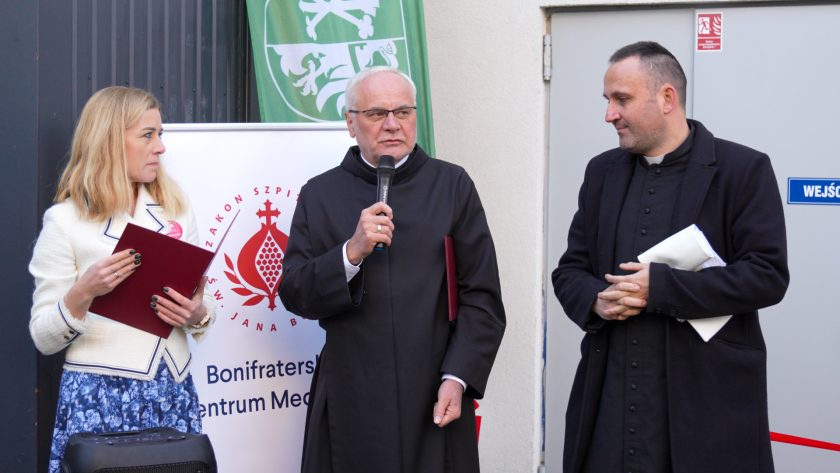 Zdjęcie przedstawia trzy osoby stojące obok siebie przed budynkiem hospicjum. Po lewej kobieta w jasnym żakiecie i niebieskiej wzorzystej spódnicy trzyma w dłoniach bordową teczkę i spogląda w stronę przemawiającego. W centrum zakonnik w czarnym habicie mówi do mikrofonu, trzymając w drugiej ręce teczkę. Po prawej stronie stoi drugi kapłan w sutannie, z dokumentami zwiniętymi w rulon. W tle widoczne są banery Bonifraterskiego Centrum Medycznego i Powiatu Średzkiego oraz wejście oznaczone tabliczką „WEJŚCIE”