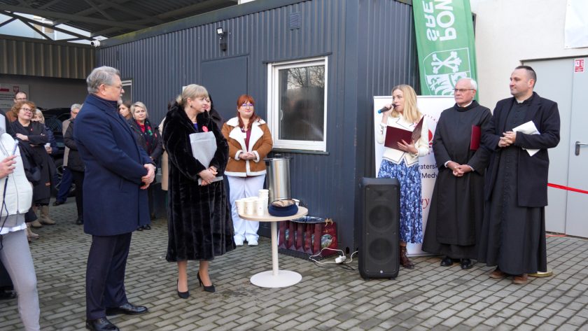 Grupa zgromadzonych osób stoi na placu przed budynkiem podczas oficjalnej ceremonii. Po prawej stronie kobieta w jasnym żakiecie przemawia do mikrofonu, trzymając bordową teczkę. Obok niej stoją dwaj zakonnicy – jeden w habicie z dokumentami w dłoni, drugi z teczką pod pachą. Po lewej stronie zdjęcia widoczni są przedstawiciele władz i uczestnicy wydarzenia, m.in. mężczyzna w granatowym płaszczu oraz kobieta w futrzanym płaszczu, trzymająca teczkę. W centrum znajduje się mały stolik z kubkami i termosami. W tle widać ścianę budynku i zielony baner Powiatu Średzkiego