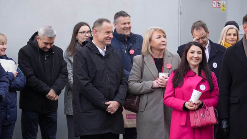 Na zdjęciu widać kilkanaście osób stojących obok siebie podczas uroczystości na świeżym powietrzu. W pierwszym rzędzie znajdują się uśmiechnięta kobieta w intensywnie różowym płaszczu, trzymająca biały kubek i różową torebkę, oraz kobieta w jasnoszarym płaszczu z przypiętą okrągłą odznaką. Obok nich stoi mężczyzna w czarnej kurtce, również z odznaką, a za nimi kilku kolejnych uczestników — część uśmiecha się, inni przysłuchują się z powagą. W tle widoczna jest szara ściana budynku i fragment drzwi technicznych. Atmosfera jest skupiona, ale pogodna