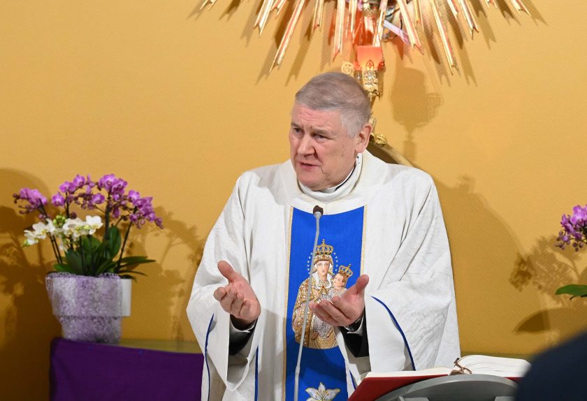 Kapłan w białych szatach liturgicznych z niebieską stułą ozdobioną wizerunkiem Maryi i Dzieciątka gestykuluje dłońmi podczas homilii. Za nim widoczna jest dekoracja ołtarzowa oraz kwiaty w doniczkach