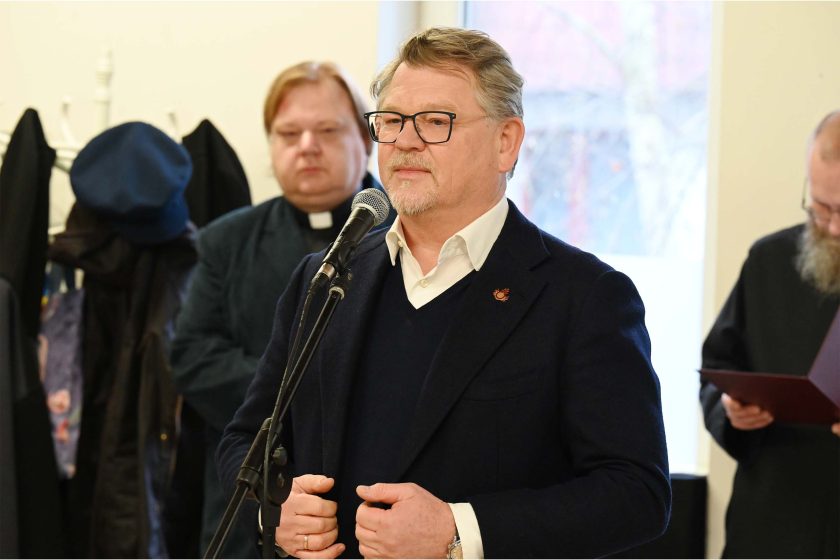 Mężczyzna w granatowej marynarce i swetrze przemawia przy mikrofonie; w tle stoją uczestnicy uroczystości, m.in. duchowny w koloratce oraz osoba w habicie z teczką