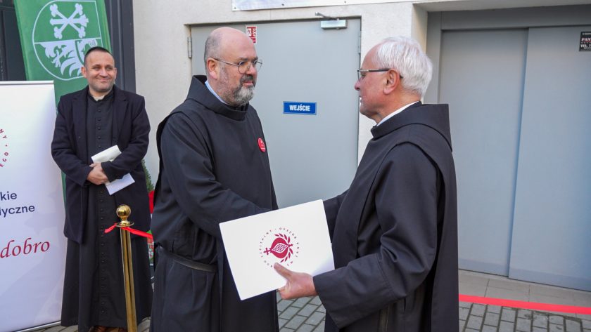 Dwóch braci zakonnych w czarnych habitach podaje sobie dłonie w geście powitania lub gratulacji. Jeden z nich trzyma biały dokument z logo Zakonu Bonifratrów. W tle widać zielony roll-up oraz fragment budynku z szarymi drzwiami