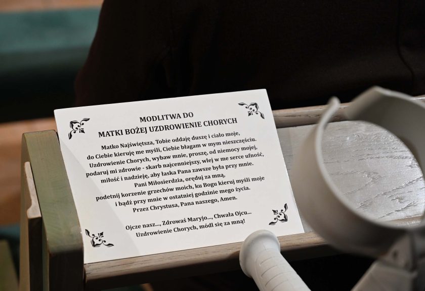 Zbliżenie na kartkę z wydrukowaną modlitwą do Matki Bożej Uzdrowienia Chorych, leżącą na oparciu ławki. Obok widać fragment kul ortopedycznych