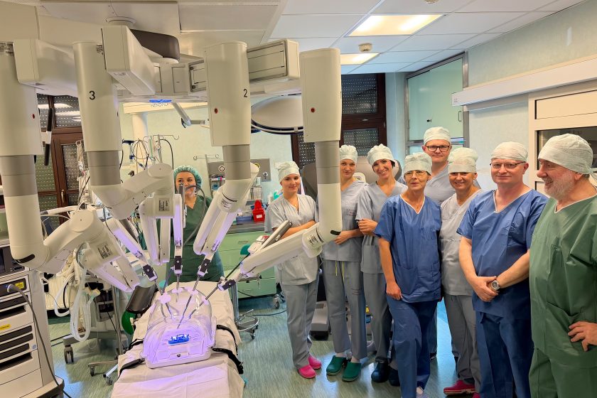 Zespół pielęgniarski, instrumentariuszki i lekarze pozują do wspólnego zdjęcia przy robocie chirurgicznym da Vinci. Ramiona robota ustawione są nad trenażerem, a tło stanowi sala operacyjna wyposażona w aparaturę medyczną. Uczestnicy uśmiechają się po zakończonym szkoleniu
