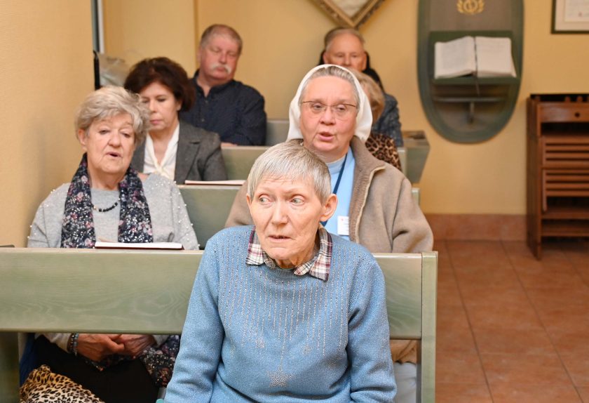 Starsze kobiety oraz siostra zakonna w białym welonie siedzą w ławkach kaplicy i modlą się lub śpiewają. Na pierwszym planie widoczna jest starsza kobieta w niebieskim swetrze z wyrazem skupienia na twarzy