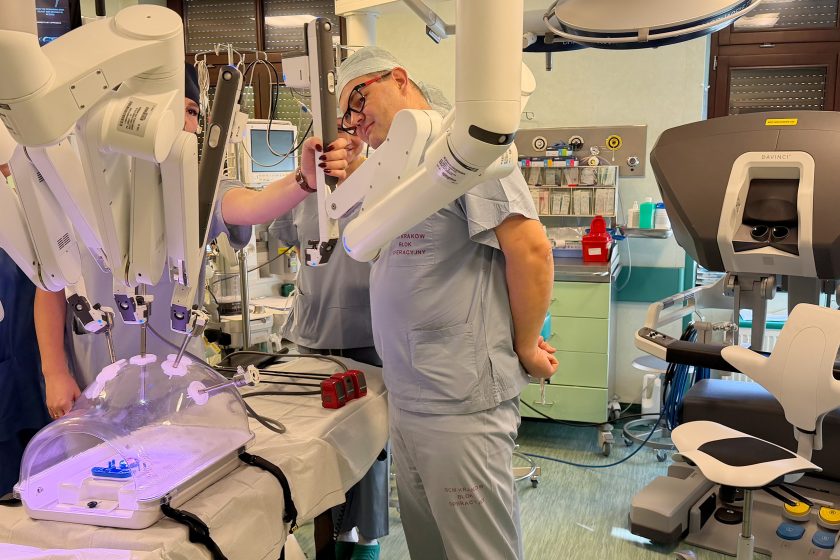 Chirurg w stroju operacyjnym pochyla się nad jednym z ramion robota da Vinci, kontrolując ustawienie narzędzia. Obok widoczna część konsoli operatorskiej oraz stół treningowy z przezroczystą kopułą. Zdjęcie ukazuje koncentrację i precyzję pracy z robotem