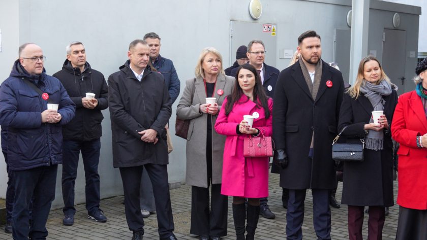 Grupa uczestników, głównie przedstawicieli lokalnych władz i instytucji, stoi przed budynkiem podczas uroczystości. Na pierwszym planie osoby ubrane elegancko, w tym kobieta w różowym płaszczu oraz mężczyzna w ciemnym płaszczu. Wszyscy patrzą w kierunku przemawiających, trzymając kubki z gorącym napojem