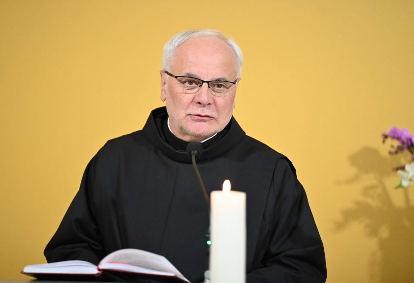 Zakonnik w czarnym habicie, w okularach, przemawia przy ambonie, trzymając otwartą księgę liturgiczną. Przed nim stoi zapalona świeca, a tło stanowi jasna ściana kaplicy