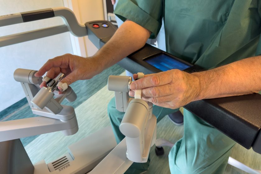 Zbliżenie na dłonie operatora obsługującego konsolę robota chirurgicznego da Vinci. Chirurg, ubrany w zielony strój operacyjny, trzyma specjalne manipulatory sterujące ramionami robota. Na palcach widoczne są zabezpieczające opaski, a w tle panel sterowania z ekranem dotykowym