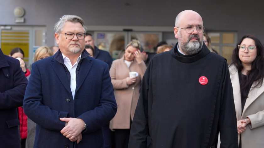 Mężczyzna w granatowym płaszczu stoi obok zakonnika w czarnym habicie. Oboje patrzą w kierunku przemawiającego. W tle widać innych uczestników, w tym kobietę poprawiającą włosy oraz osoby z papierowymi kubkami