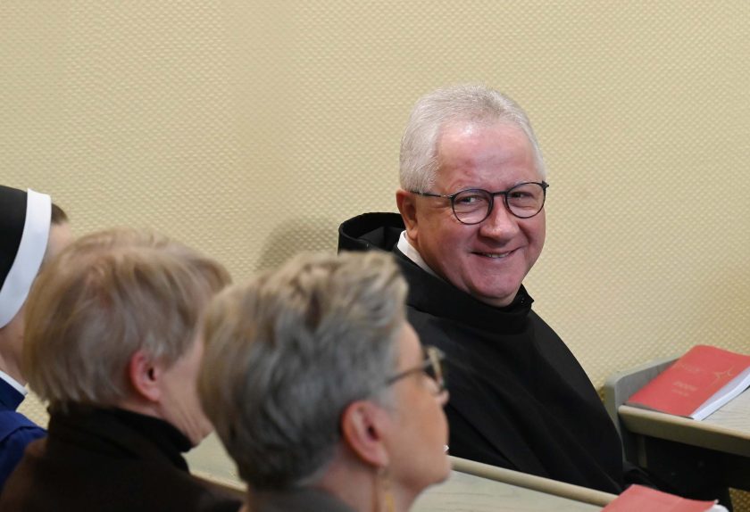 Zakonnik w czarnym habicie siedzi w ławce kaplicy i uśmiecha się w stronę fotografa. Wokół niego siedzą świeckie kobiety oraz siostra zakonna, a na ławkach leżą czerwone modlitewniki