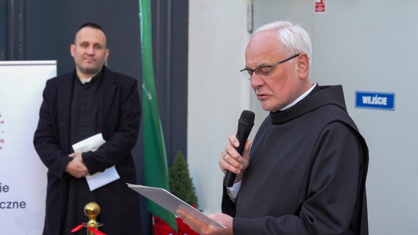Brat zakonny w czarnym habicie czyta przemówienie z kartki, trzymając mikrofon w ręku. Obok, po lewej stronie, stoi inny duchowny ubrany w czarną sutannę, trzymający dokumenty. Za nimi znajduje się roll-up z logo Bonifraterskiego Centrum Medycznego oraz zielona flaga