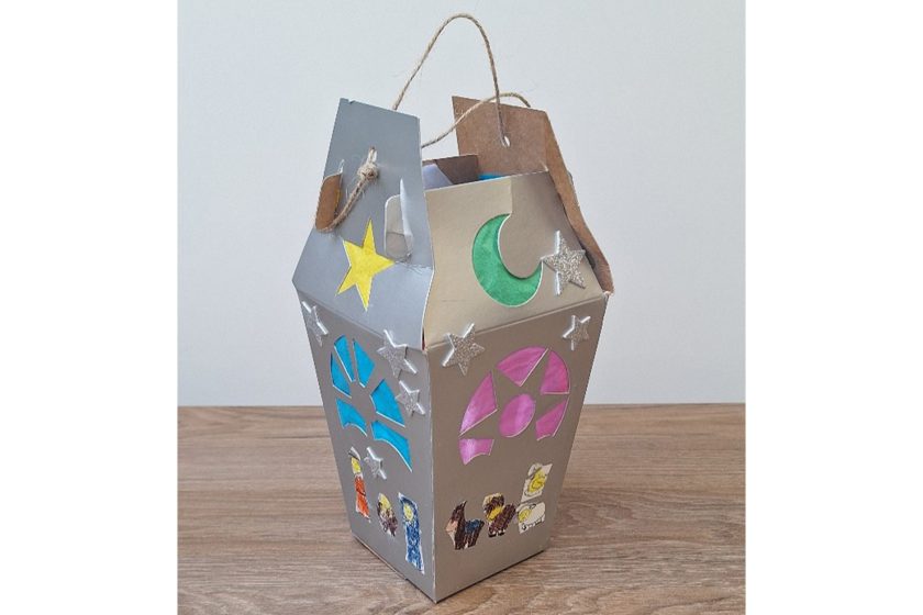 Kartonowy lampion w formie domku, dekorowany gwiazdkami, półksiężycem i kolorowymi witrażowymi wycięciami, stojący na drewnianej powierzchni