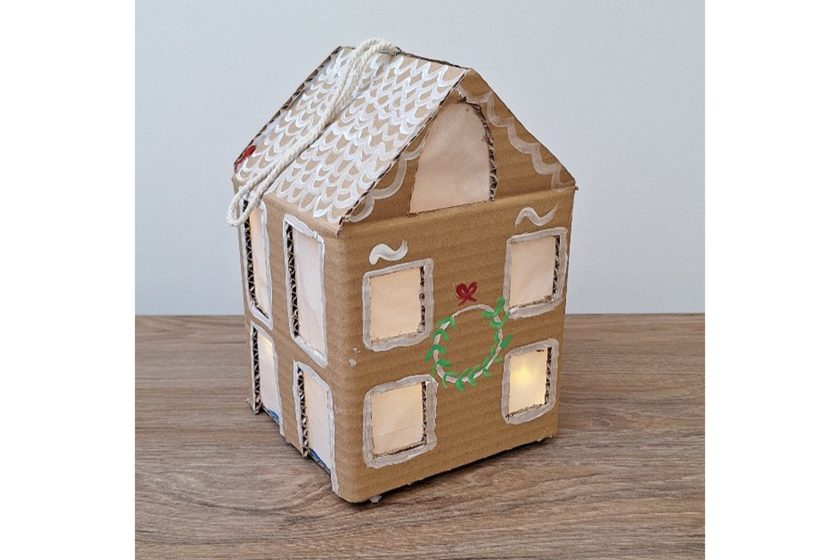 Kartonowy domek z papieru, z wyciętymi oknami i białymi zdobieniami na dachu, podświetlony od środka i ustawiony na drewnianym blacie