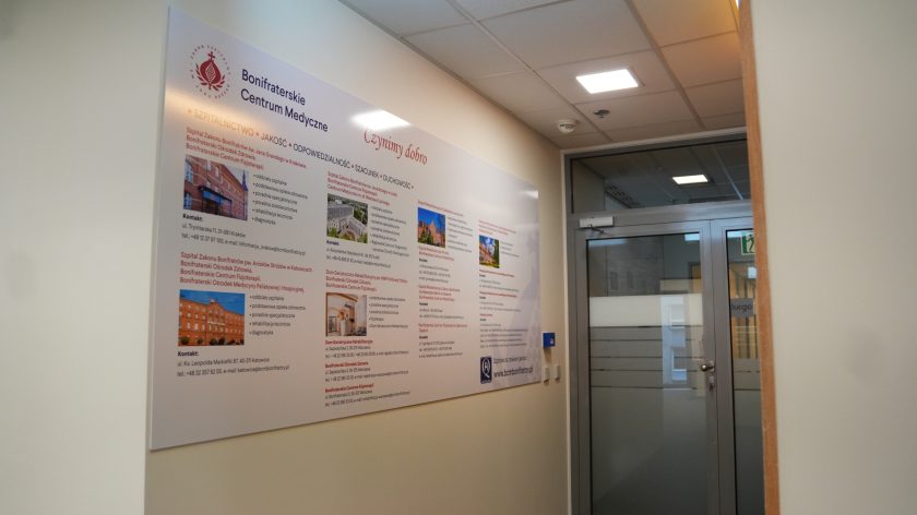 Korytarz w budynku Bonifraterskiego Centrum Medycznego. Po lewej stronie duża plansza informacyjna przedstawiająca oddziały, placówki i działalność Zakonu Bonifratrów. W głębi widoczne przeszklone drzwi wejściowe oraz oznaczenia kierunkowe