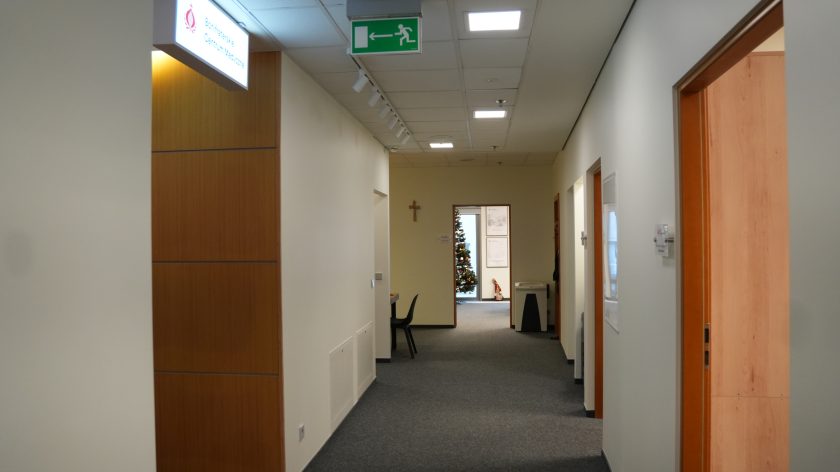 Jasny, długi korytarz biurowy w Bonifraterskim Centrum Medycznym. Po lewej stronie pod sufitem podświetlany szyld z nazwą placówki, wyżej znak ewakuacyjny. W głębi widoczne pomieszczenie z choinką świąteczną i krzyżem zawieszonym na ścianie