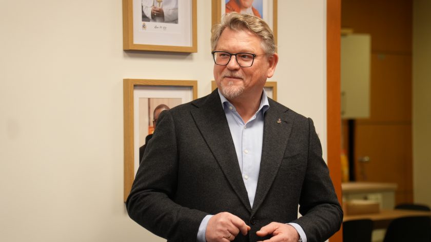 Mężczyzna w eleganckiej marynarce stoi w biurze, uśmiechając się. Za nim na ścianie wiszą w ramkach portrety duchownych. W tle widoczne są elementy wyposażenia biura