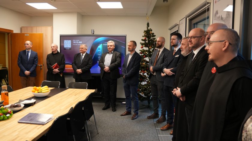 Grupa mężczyzn – w tym świeccy oraz zakonnicy w czarnych habitach – stoi w sali konferencyjnej podczas uroczystości. W tle widoczna jest choinka świąteczna oraz duży ekran multimedialny. Na stole przed nimi leżą owoce, napoje i przekąski