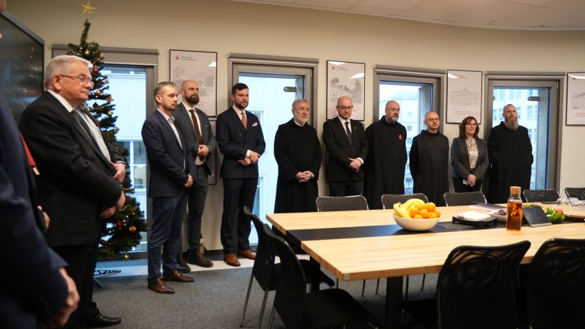 Kilkanaście osób stoi wzdłuż ściany sali konferencyjnej, część w garniturach, część w habitach. W tle widoczne są ramki z rysunkami budynków Bonifratrów oraz okna. Przy stole ustawione są owoce i poczęstunek przygotowany na spotkanie