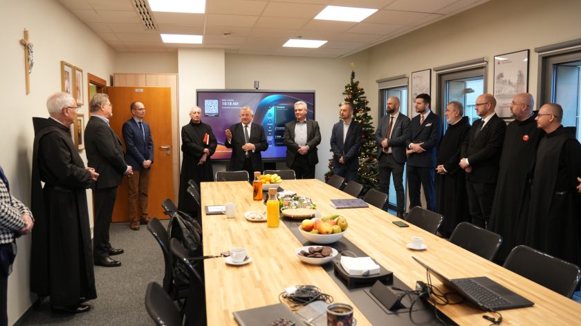 Grupa mężczyzn i zakonników stoi w sali konferencyjnej tworząc półokrąg wokół stołu. Przy stole leżą owoce, ciastka, dzbanek soku oraz dokumenty. W tle ekran multimedialny wyświetla godzinę i datę, obok stoi świąteczna choinka