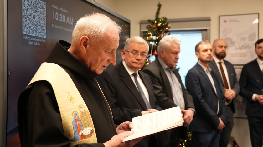 Zakonnik w habicie i kremowej stule czyta fragment modlitwy lub błogosławieństwa z książki. Obok niego stoi kilku mężczyzn w garniturach oraz zakonnicy. W tle widać choinkę świąteczną oraz ekran multimedialny