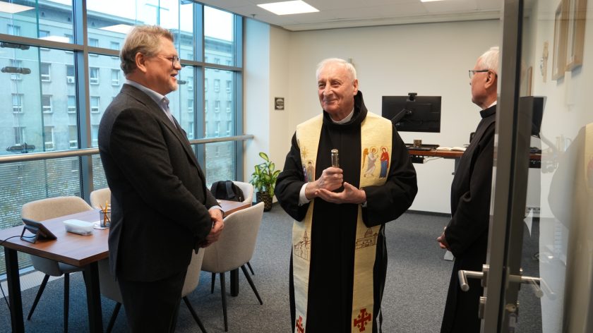 W jasnym biurze trzech mężczyzn stoi i rozmawia. Po lewej stronie mężczyzna w garniturze uśmiecha się, zwrócony w stronę zakonnika. W centrum stoi zakonnik w czarnym habicie i kremowej stule z wizerunkami świętych, trzymający w dłoniach pojemnik z wodą święconą. Po prawej kolejny zakonnik słucha rozmowy. W tle widać duże okna oraz biurko z krzesłami