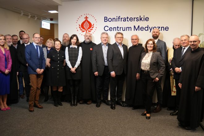 Zdjęcie grupowe wykonane na tle ściany z logo Zakonu Szpitalnego św. Jana Bożego i napisem ‘Bonifraterskie Centrum Medyczne’. W jednym rzędzie stoi kilkanaście osób – świeccy pracownicy w garniturach i eleganckich strojach oraz zakonnicy w czarnych habitach. Uczestnicy patrzą w stronę obiektywu i uśmiechają się. Po lewej stronie widoczny fragment korytarza, po prawej zielona roślina w donicy ustawionej na szafce