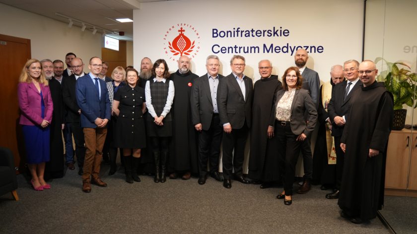 Zdjęcie grupowe wykonane na tle ściany z logo Zakonu Szpitalnego św. Jana Bożego i napisem ‘Bonifraterskie Centrum Medyczne’. W jednym rzędzie stoi kilkanaście osób – świeccy pracownicy w garniturach i eleganckich strojach oraz zakonnicy w czarnych habitach. Uczestnicy patrzą w stronę obiektywu i uśmiechają się. Po lewej stronie widoczny fragment korytarza, po prawej zielona roślina w donicy ustawionej na szafce